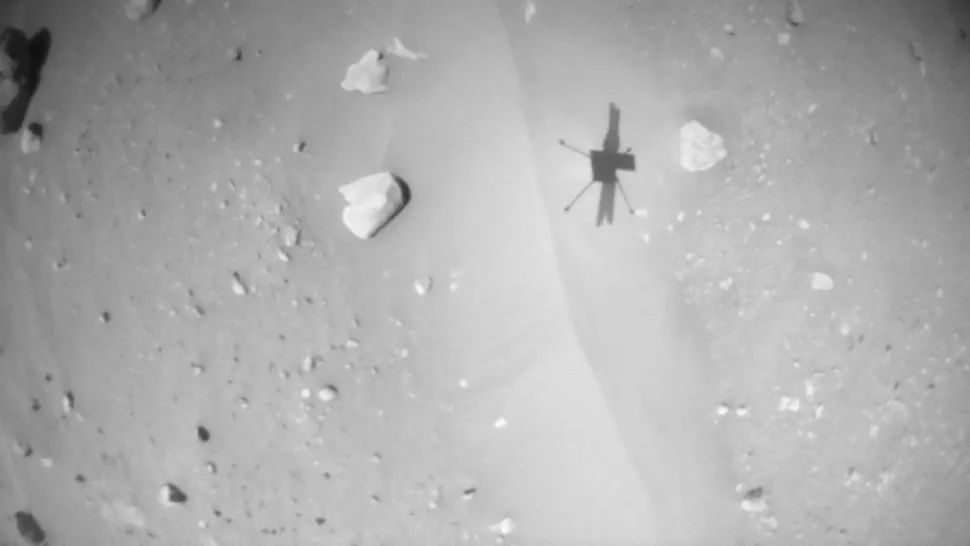een zwart-witafbeelding die de schaduw van een kleine helikopter laat zien op een zanderige, met keien bezaaide grond.